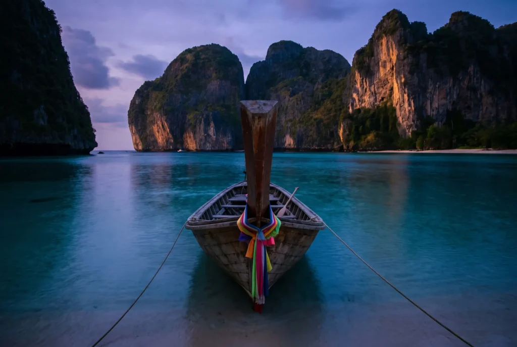 An empty longtail boat moored in shallow water at dusk below the Railay cliffs, the only route off the peninsula